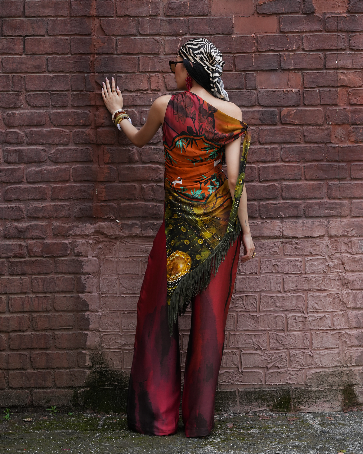 A woman stands with her back to the camera against a red brick wall, wearing a colorful draped dress with a zebra-print headscarf and gold bracelets.