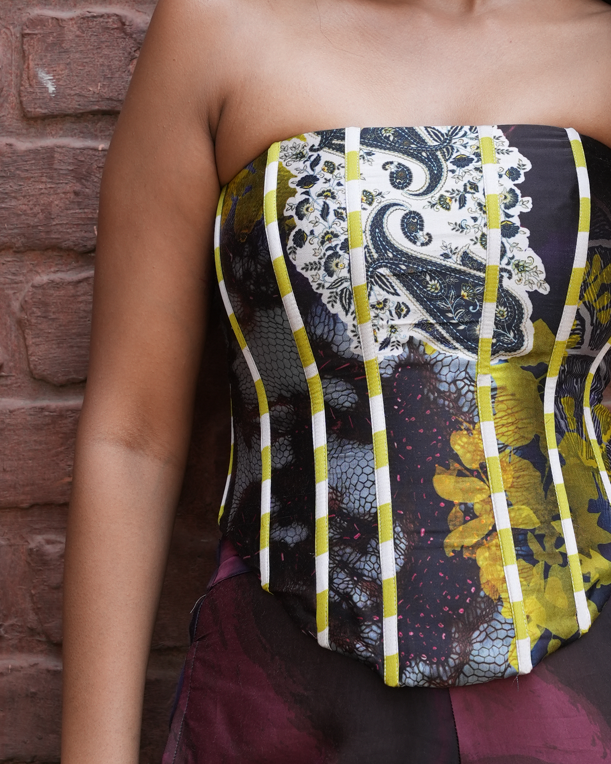 A close-up of a model wearing a strapless corset-style top with a complex abstract print featuring yellow, black, white, and gray, standing against a red-brown brick wall.