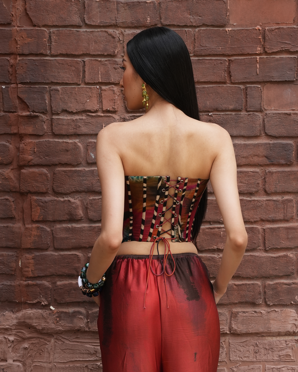 A woman is shown from behind wearing a strapless corset-style top with vertical patterned panels and red satin-like pants, standing against a red-brown brick wall.
