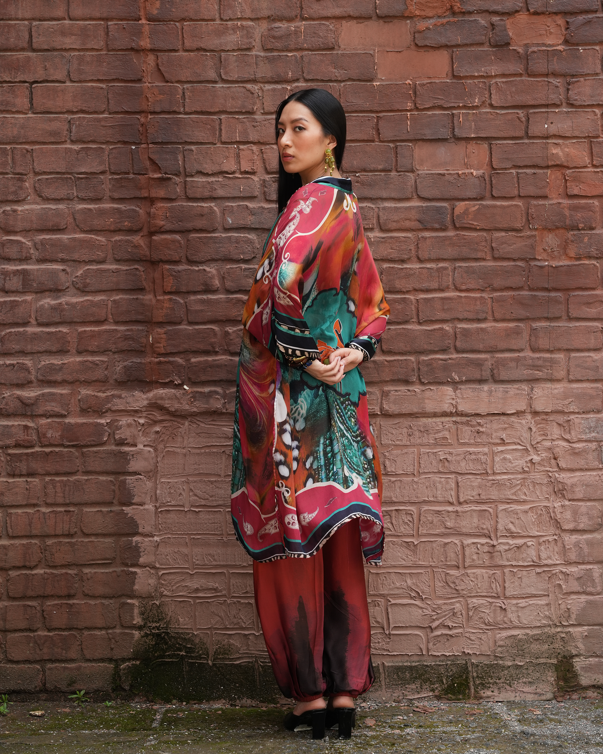 A woman stands in profile against a weathered red-brown brick wall, wearing a colorful printed traditional outfit with a long draped tunic, patterned skirt, and black heels.