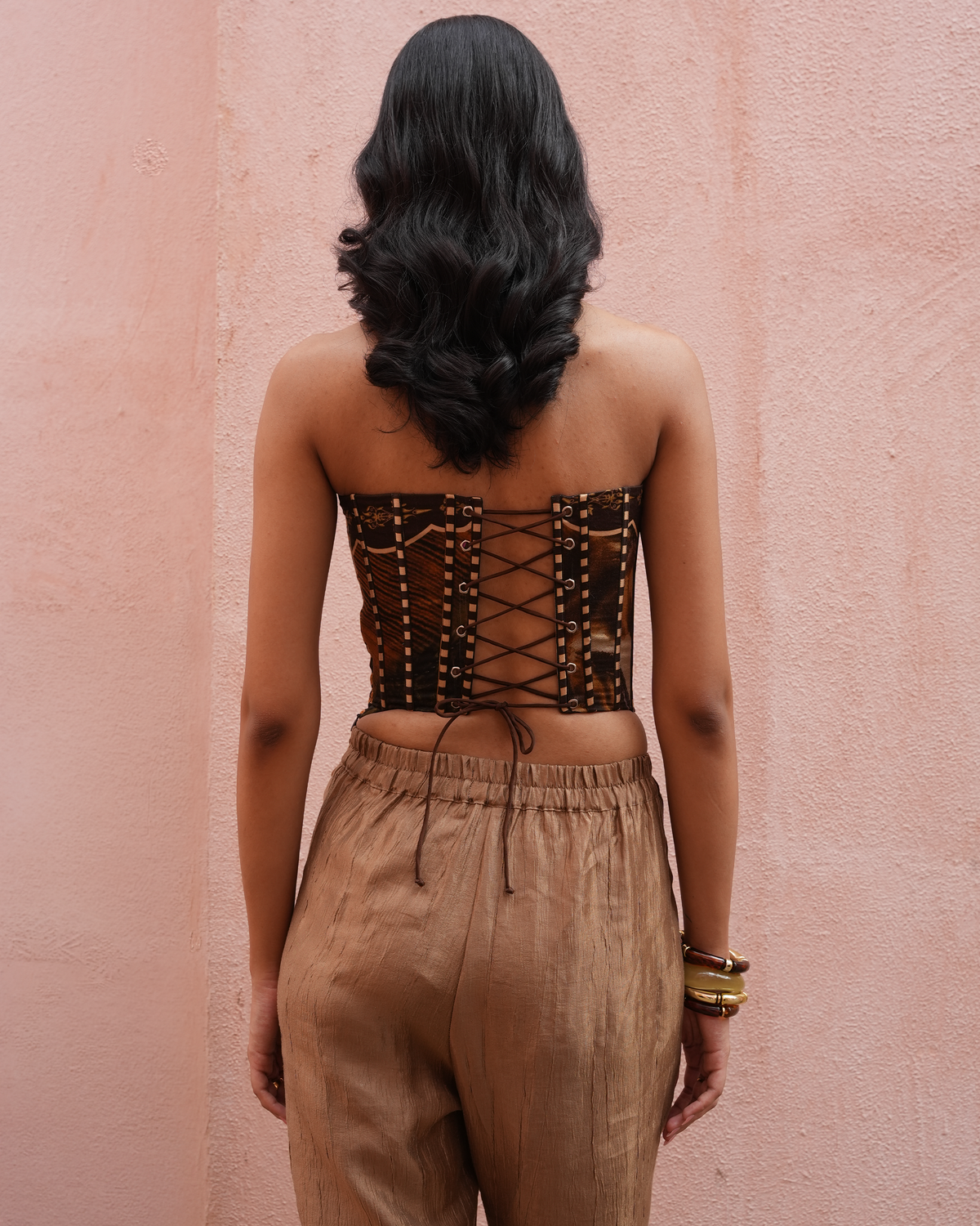 A woman viewed from behind wearing a brown strapless lace-up corset top and tan drawstring pants, standing against a textured pink wall.