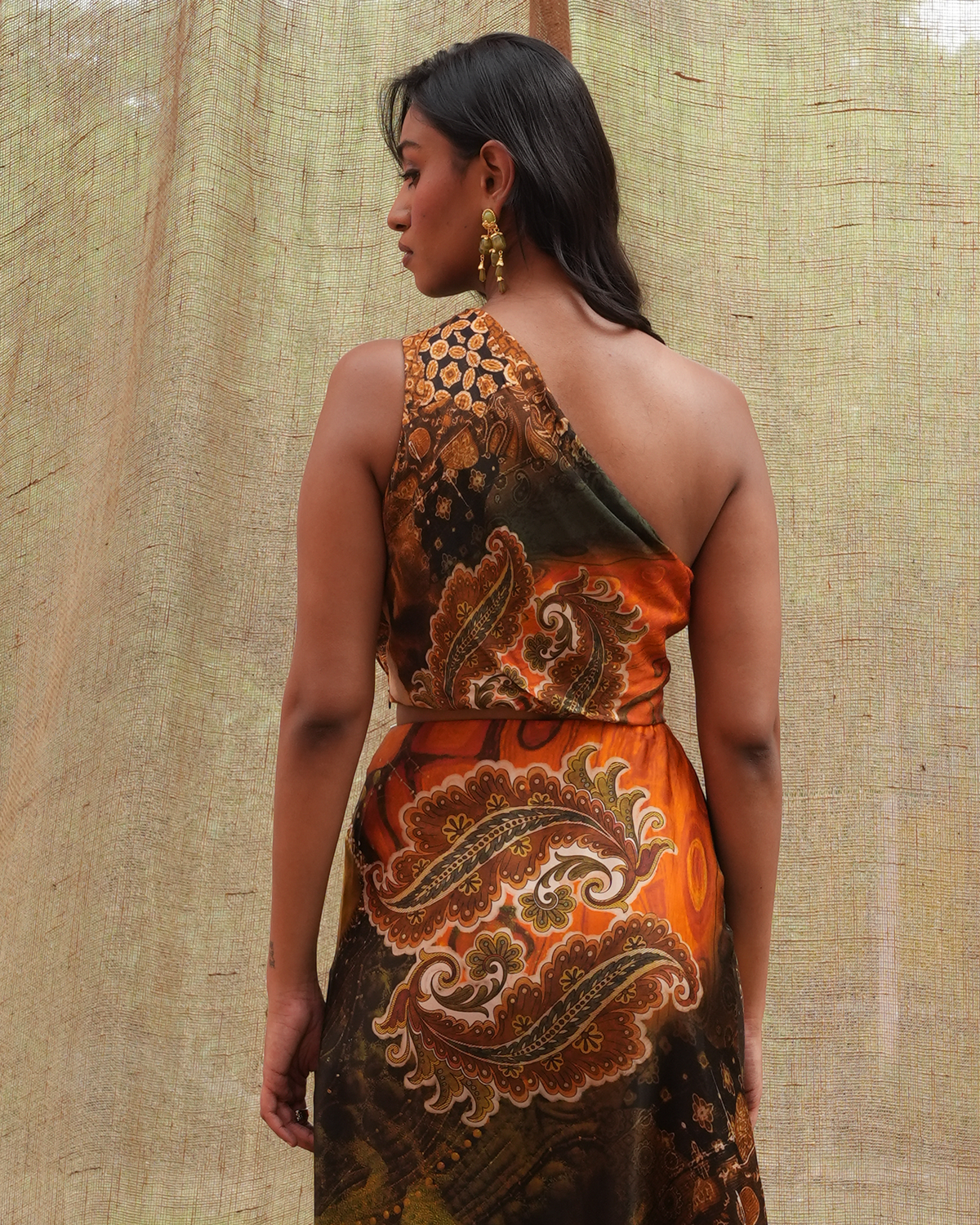 A woman shown from the back wearing a one-shoulder dress with a bold paisley print in orange, brown, and black, posed against a textured beige fabric backdrop.