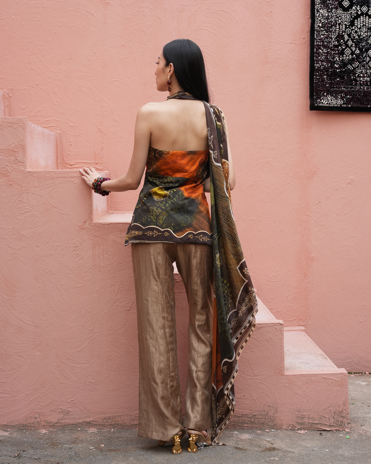 A woman stands with her back to the camera wearing a sleeveless top and wide-leg trousers in a rust, orange, and green abstract print, paired with metallic bronze wide-leg pants and a matching patterned dupatta, set against a textured pink wall.