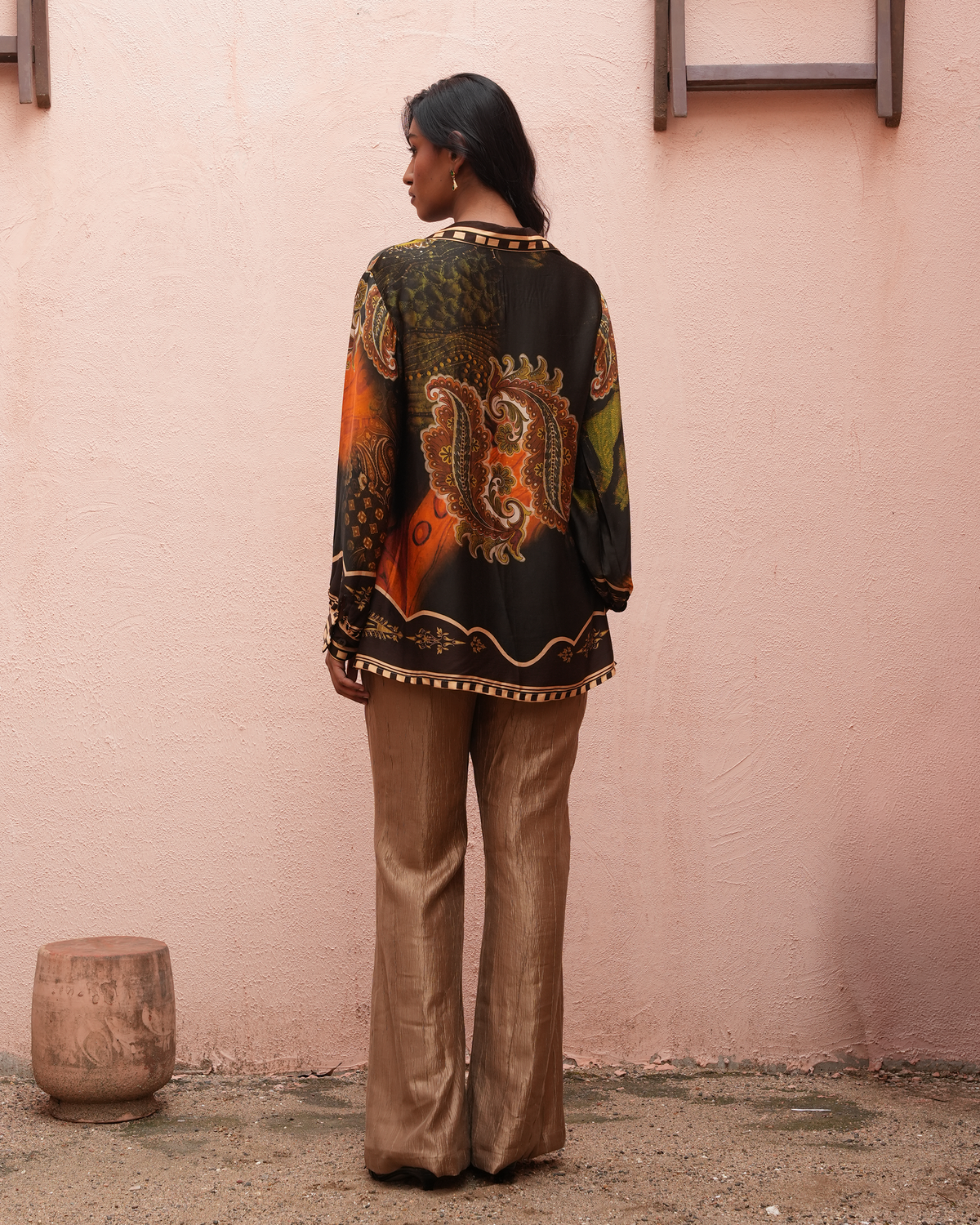 A woman stands with her back to the camera wearing a black long-sleeve blouse with a colorful paisley and floral print in orange, green, and gold, paired with metallic bronze flared pants, set against a textured pink wall.