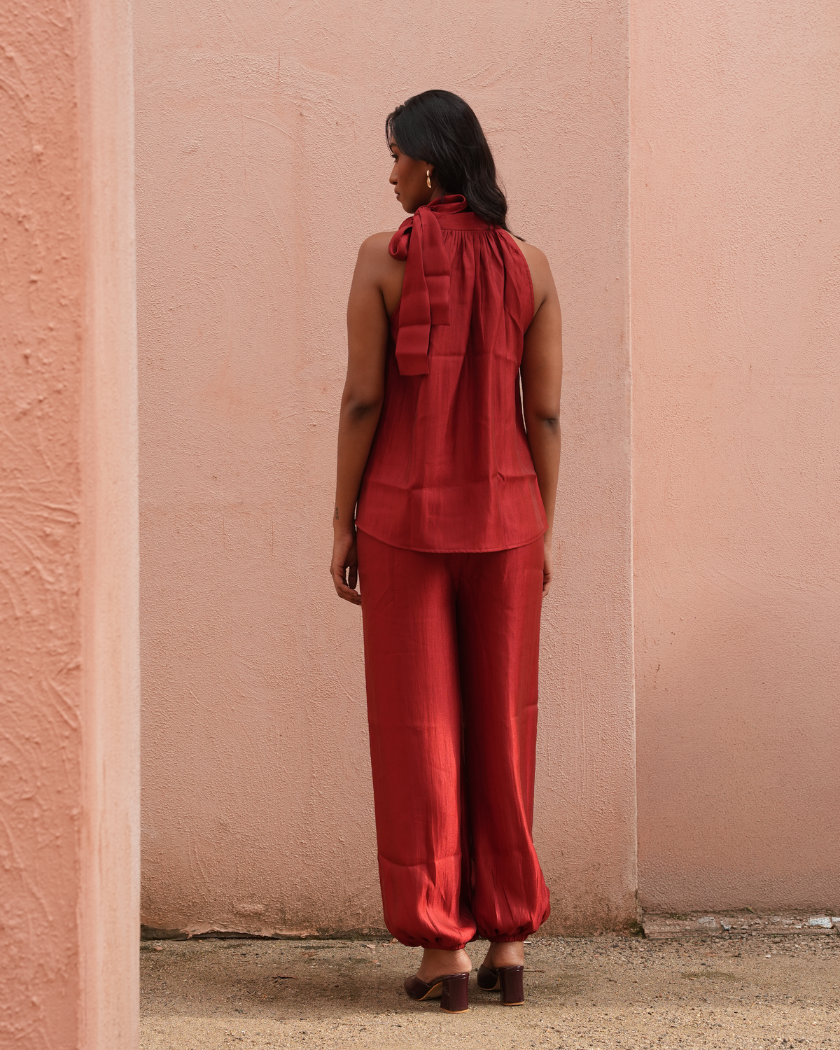 A woman stands with her back to the camera wearing a matching rust-red sleeveless top and wide-leg pants set, posed against a textured pink stucco wall.
