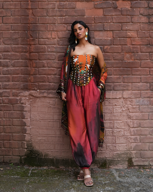 A woman stands against a red-brown brick wall wearing a strapless orange and red printed outfit with a matching draped scarf and sandals.