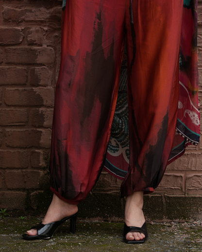 A person wearing loose, flowing pants in a deep red and black abstract print, paired with black heeled sandals, standing against a rustic brick wall.
