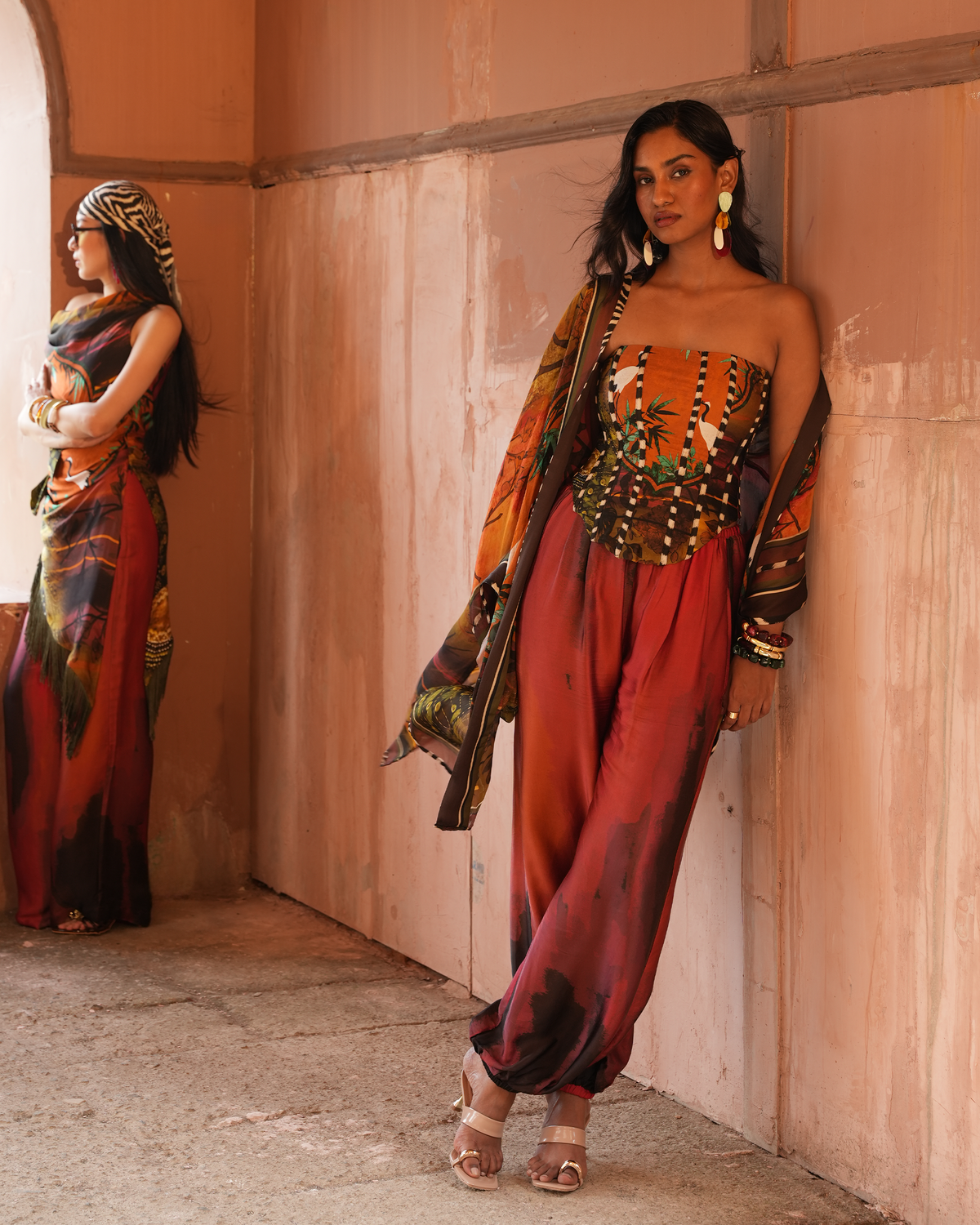 A woman models a strapless jumpsuit with a heavily embellished corset-style bodice and a draped dupatta, standing against a warm terracotta wall. A second woman in a matching printed saree stands in the background in a sunlit corridor.