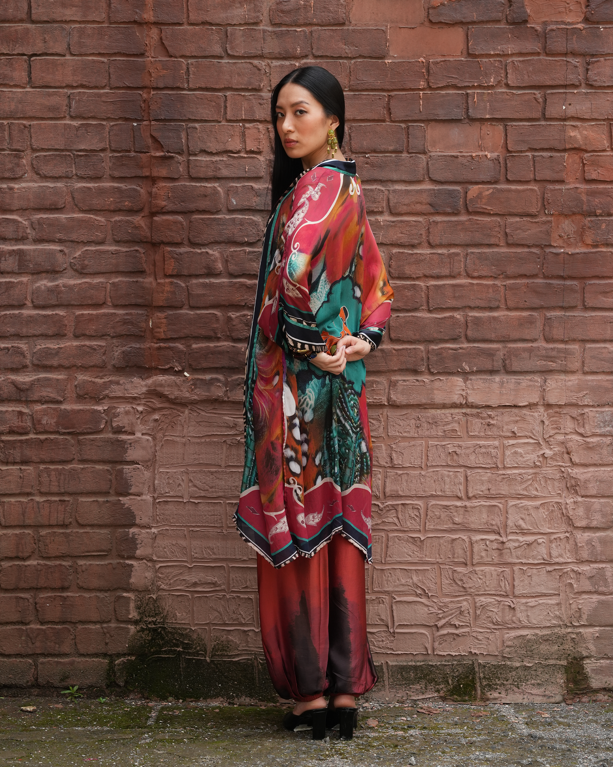 A woman stands in profile against a red-brown brick wall, wearing a long, colorful printed tunic or kaftan with wide sleeves and a wide patterned belt, paired with matching pleated skirt or pants in red and teal tones, and black pointed shoes.