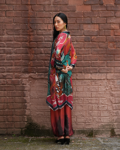 A woman stands in profile against a red-brown brick wall, wearing a long, colorful printed tunic or kaftan with wide sleeves and a wide patterned belt, paired with matching pleated skirt or pants in red and teal tones, and black pointed shoes.