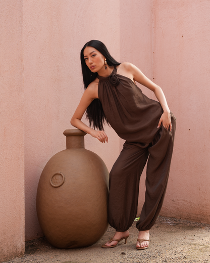A woman poses in a brown halter top and matching wide-leg pants next to a large terracotta vase against a pink stucco wall.