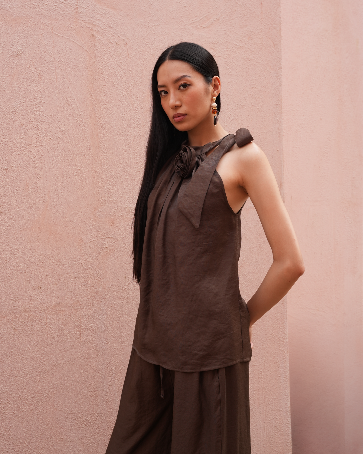 A woman with long black hair models a dark brown linen sleeveless top with a tie-neck detail and matching wide-leg trousers, standing against a textured pink wall.