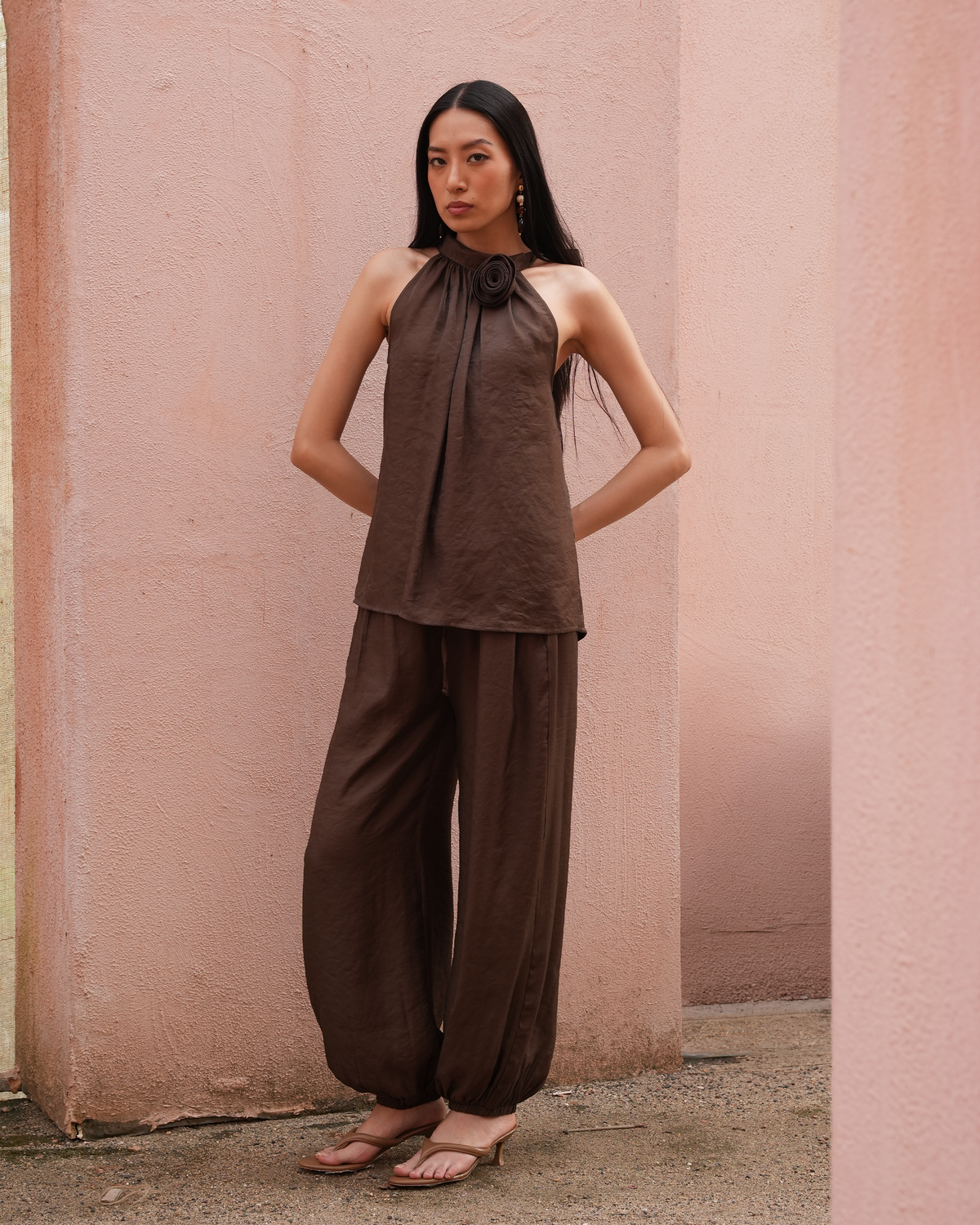 A woman modeling a dark brown two-piece linen set consisting of a halter top with a large flower detail at the collar and matching wide-leg pants, standing against a textured pink wall.