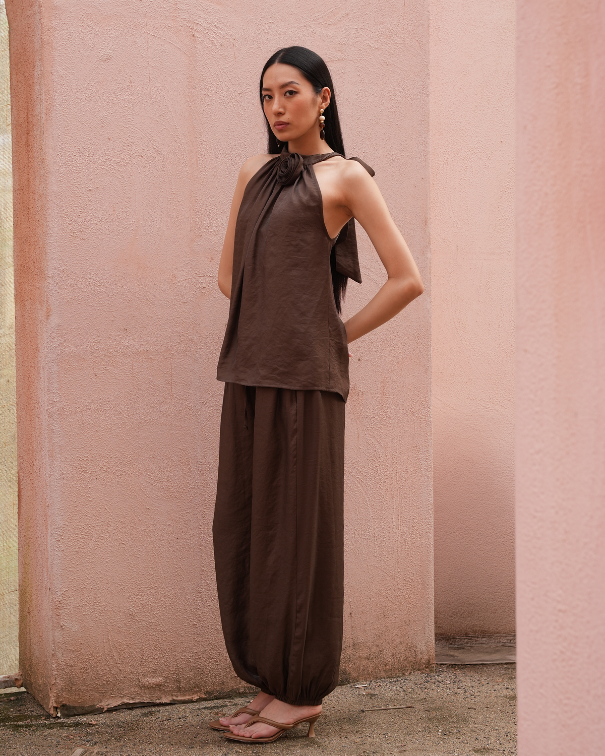 A woman stands in a real architectural setting with pink walls, wearing a matching brown halter top and loose wide-leg pants, with nude heeled sandals.