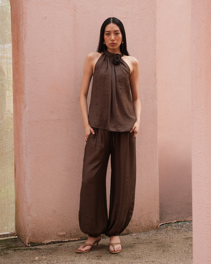 A woman stands outdoors against a pink stucco wall wearing a matching brown sleeveless halter top with a large flower detail at the neck and wide-leg brown pants.