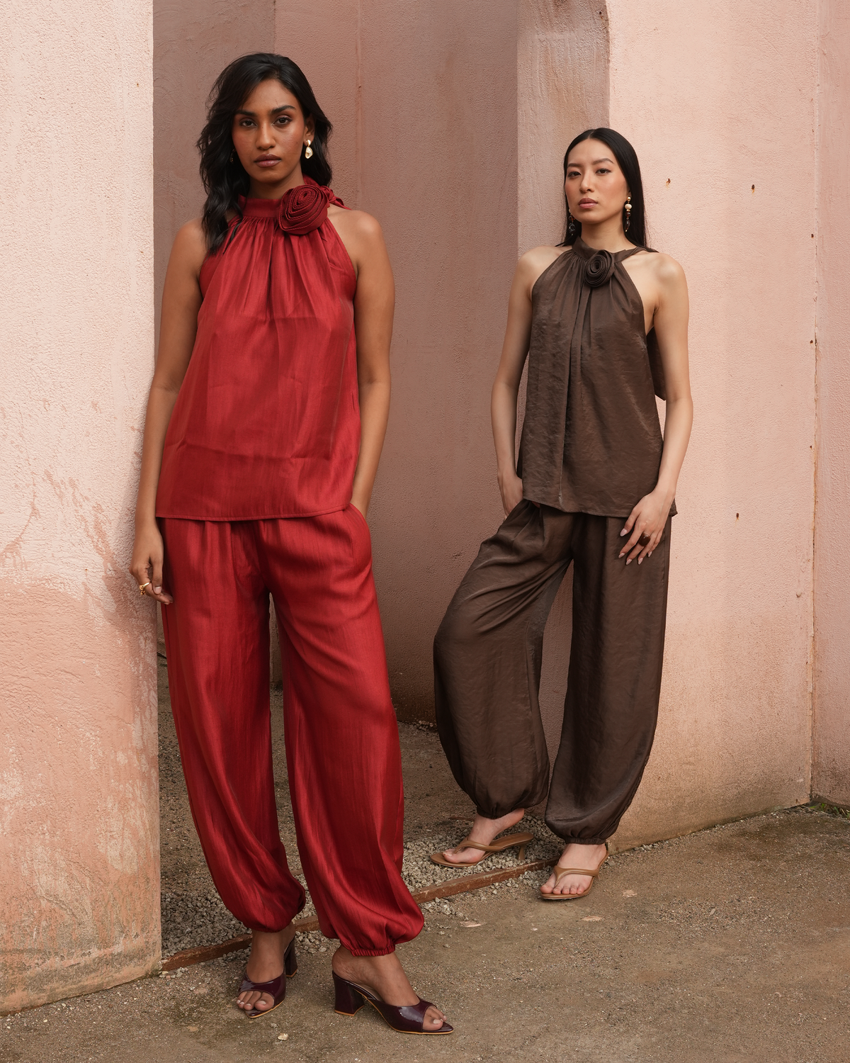 Two women modeling matching satin-like harem pants and halter tops in contrasting red and brown, standing against a textured pinkish-beige wall.