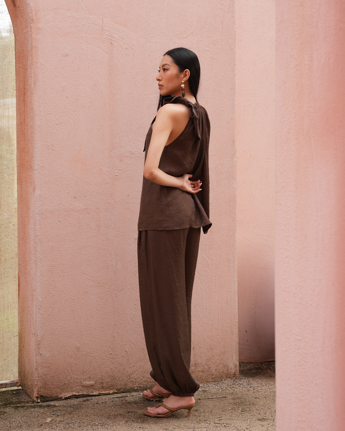 A woman stands in profile wearing a dark brown sleeveless top and matching wide-leg pants against a textured pink wall.