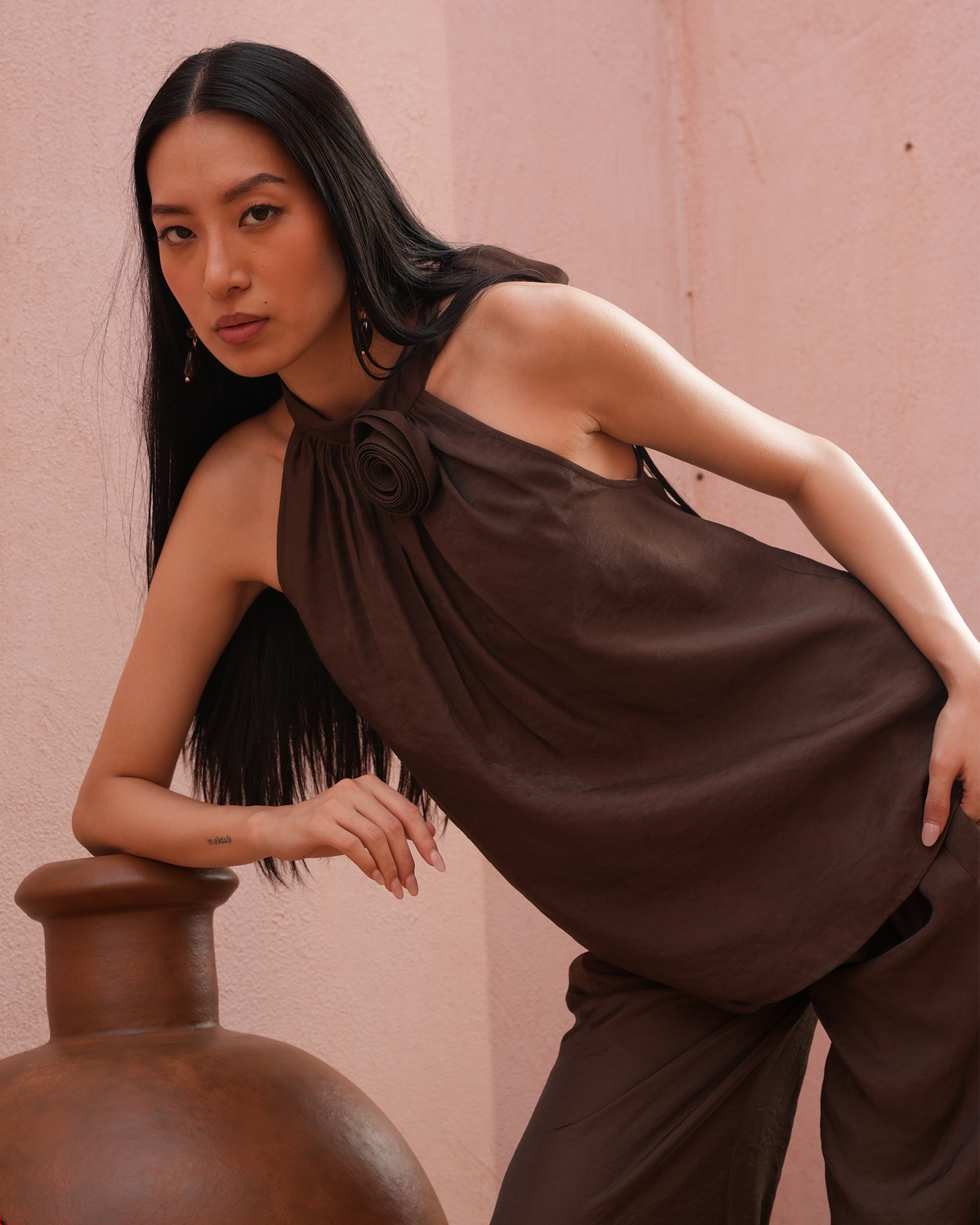 A woman with long black hair models a dark brown draped halter top and matching wide-leg trousers against a textured pink wall, leaning on a large terracotta pot.