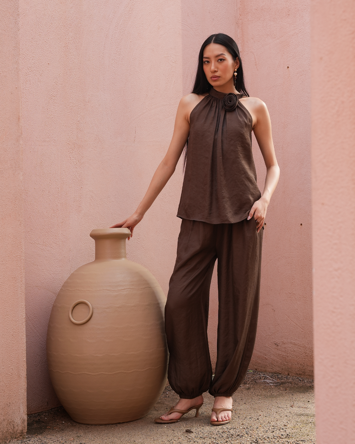 A woman stands in a pink architectural alcove wearing a dark brown sleeveless halter top and matching loose-fitting pants, posing next to a large beige ceramic vase.