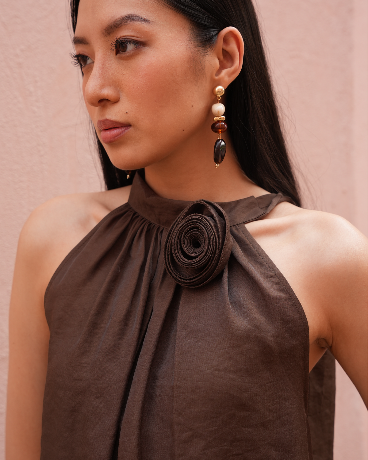 A close-up of a woman wearing a dark brown halter top with a large rolled fabric detail at the neck, paired with dangling earrings against a soft pink wall.