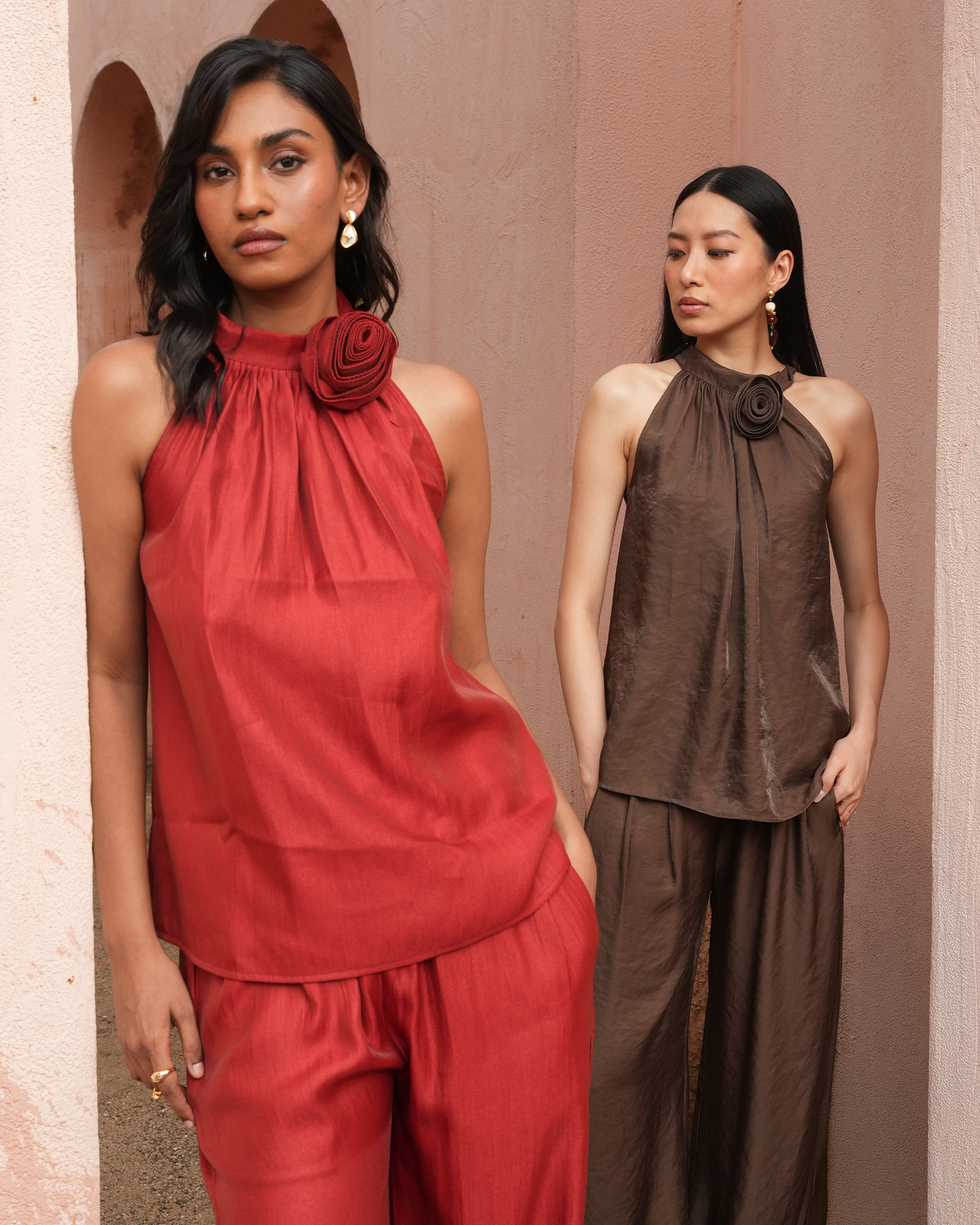 Two models wearing sleeveless halter tops and matching wide-leg pants in contrasting rust red and dark brown, styled with large fabric rose embellishments at the collar, set against a textured stucco wall.