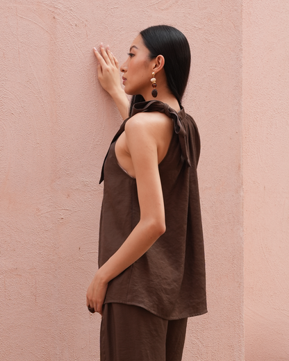 A woman in a side profile wearing a brown sleeveless top and matching pants, posing against a textured pink wall.