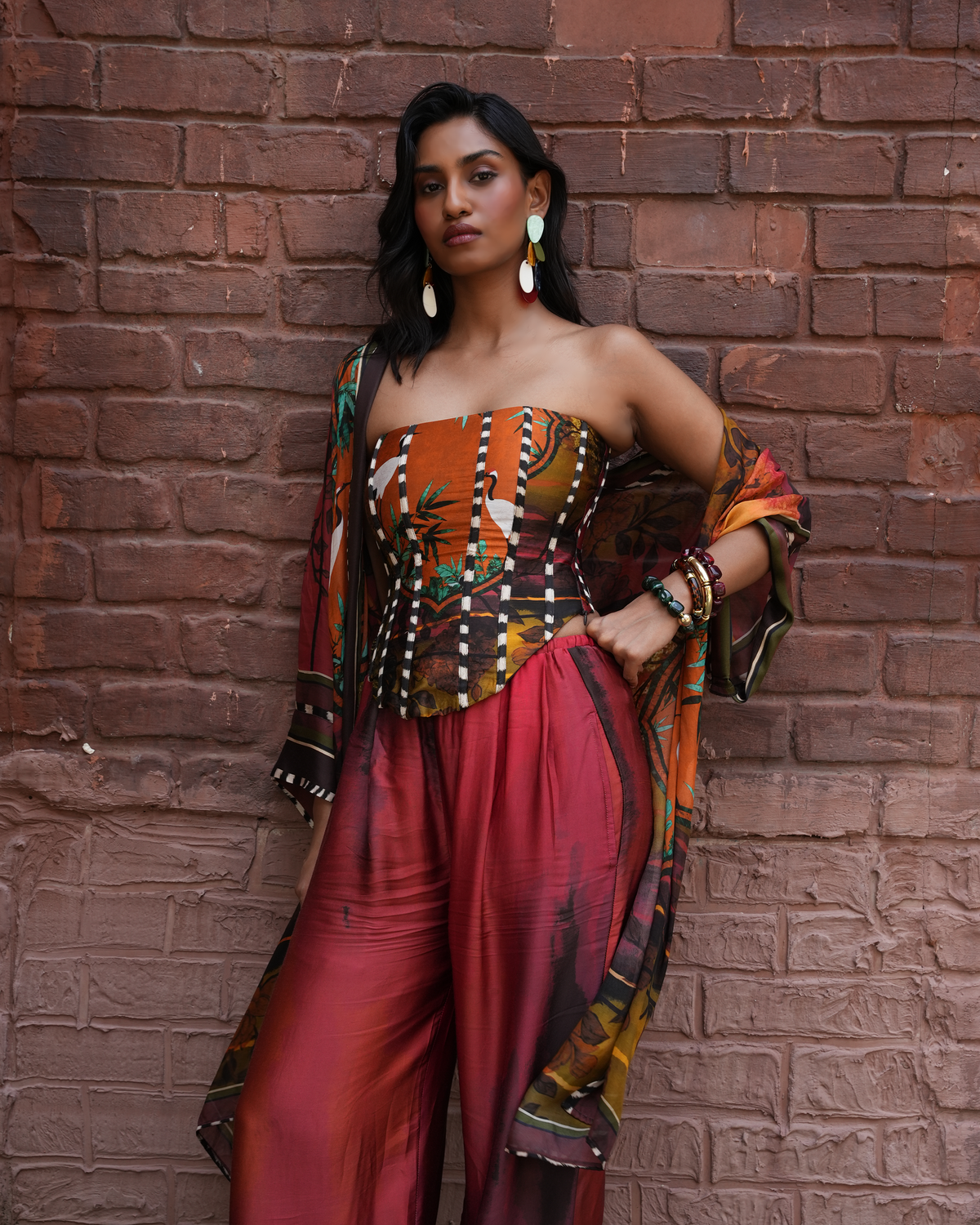 A woman posing against a red brick wall wearing a strapless jumpsuit with a multicolor abstract print in orange, brown, and red, paired with a matching draped shawl.