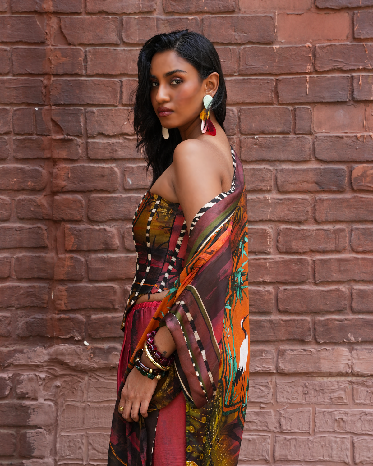 A woman posing in front of a red brick wall, wearing a colorful, patterned draped dress or sari with brown, orange, green, and red tones, accessorized with large white and red earrings and multiple gold bangles.