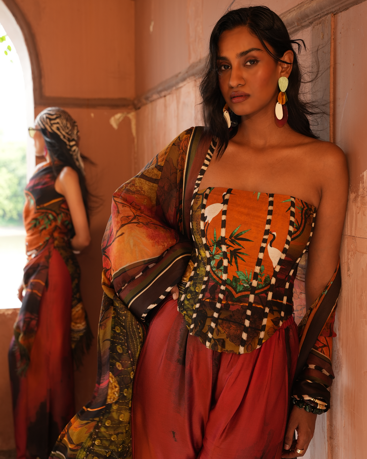 A woman models a vibrant, patterned strapless top with a matching draped scarf and rust-red pants, posing in a warm, earth-toned architectural setting with a mirror reflecting her back.