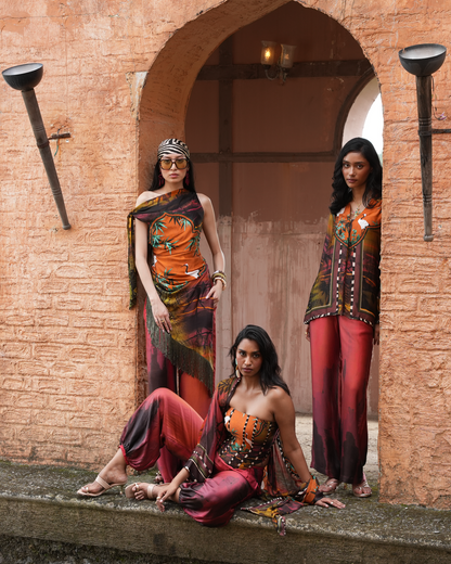 Three women modeling colorful printed ethnic wear in front of a rustic terracotta archway. The outfits feature orange, red, and dark brown tones with intricate patterns and embellishments.