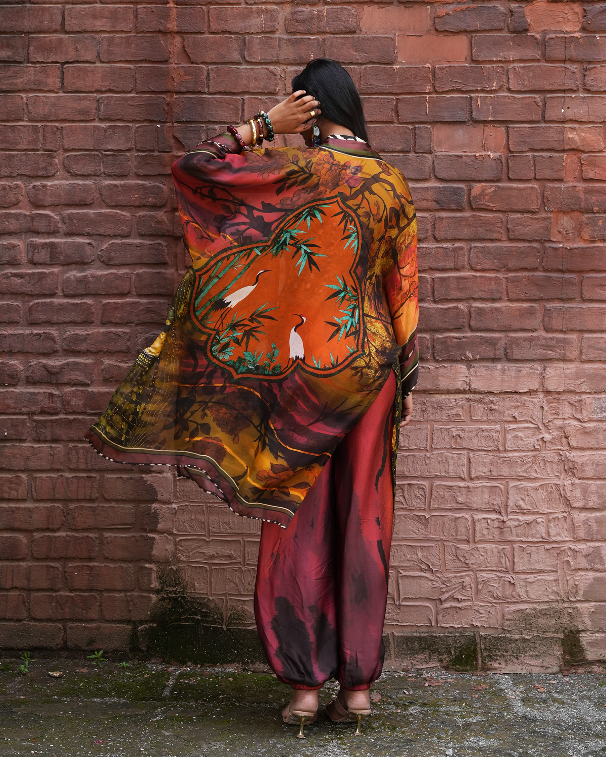 A woman stands with her back to the camera in front of a red brick wall, wearing a flowing printed shawl draped over her shoulders and matching maroon pants.