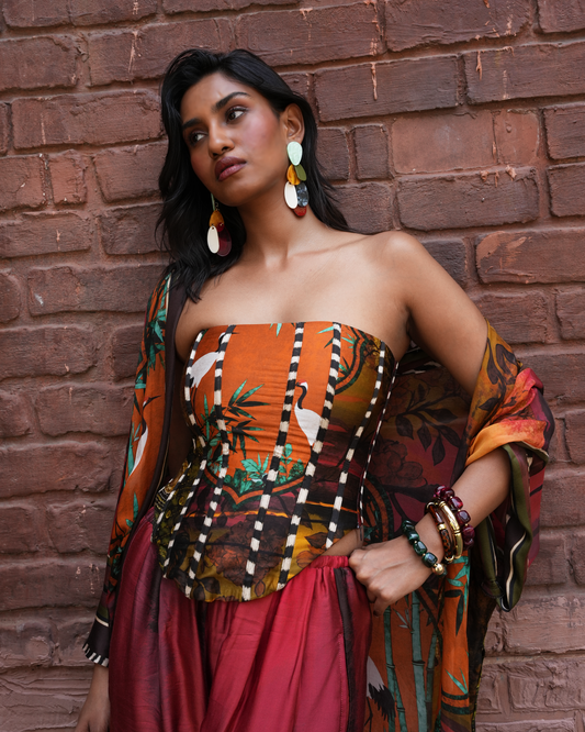 A woman posing against a rustic red brick wall wearing a strapless tube top with a tropical bird print, a matching sheer shawl, and a red skirt. She is accessorized with large multi-colored disc earrings and multiple bangles.