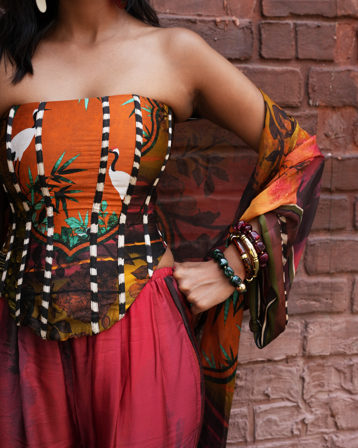 A woman wearing a strapless tube top with a vibrant orange, black, and white tropical print, paired with a matching sheer draped sleeve and a pink skirt, standing against a red brick wall.
