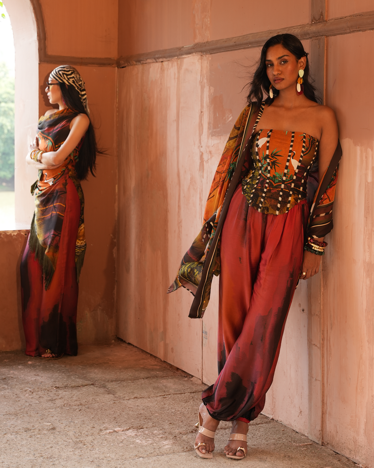 A woman models a colorful printed outfit consisting of a strapless top, wide-leg pants, and a draped shawl, standing in a warm-toned architectural setting. Another view of the same outfit is shown on a mannequin or model in the background archway.