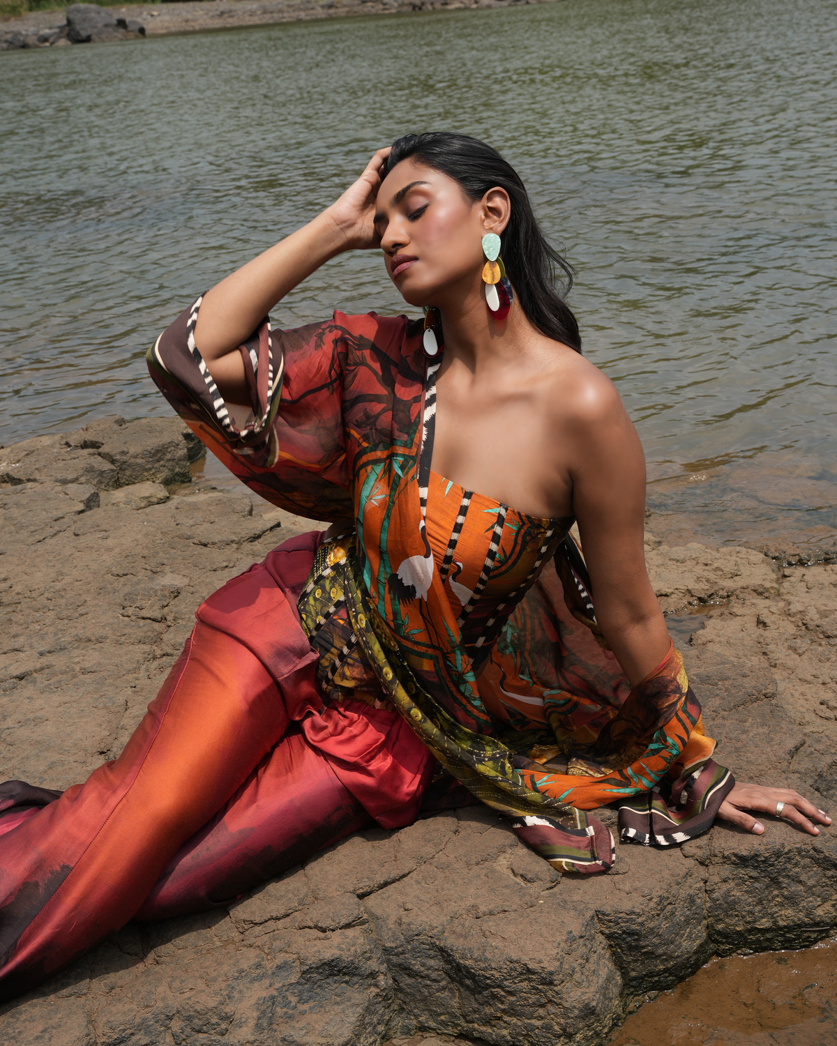 A woman models a vibrant orange and red patterned two-piece set consisting of a strapless top and matching pants, sitting on rocks next to a body of water.