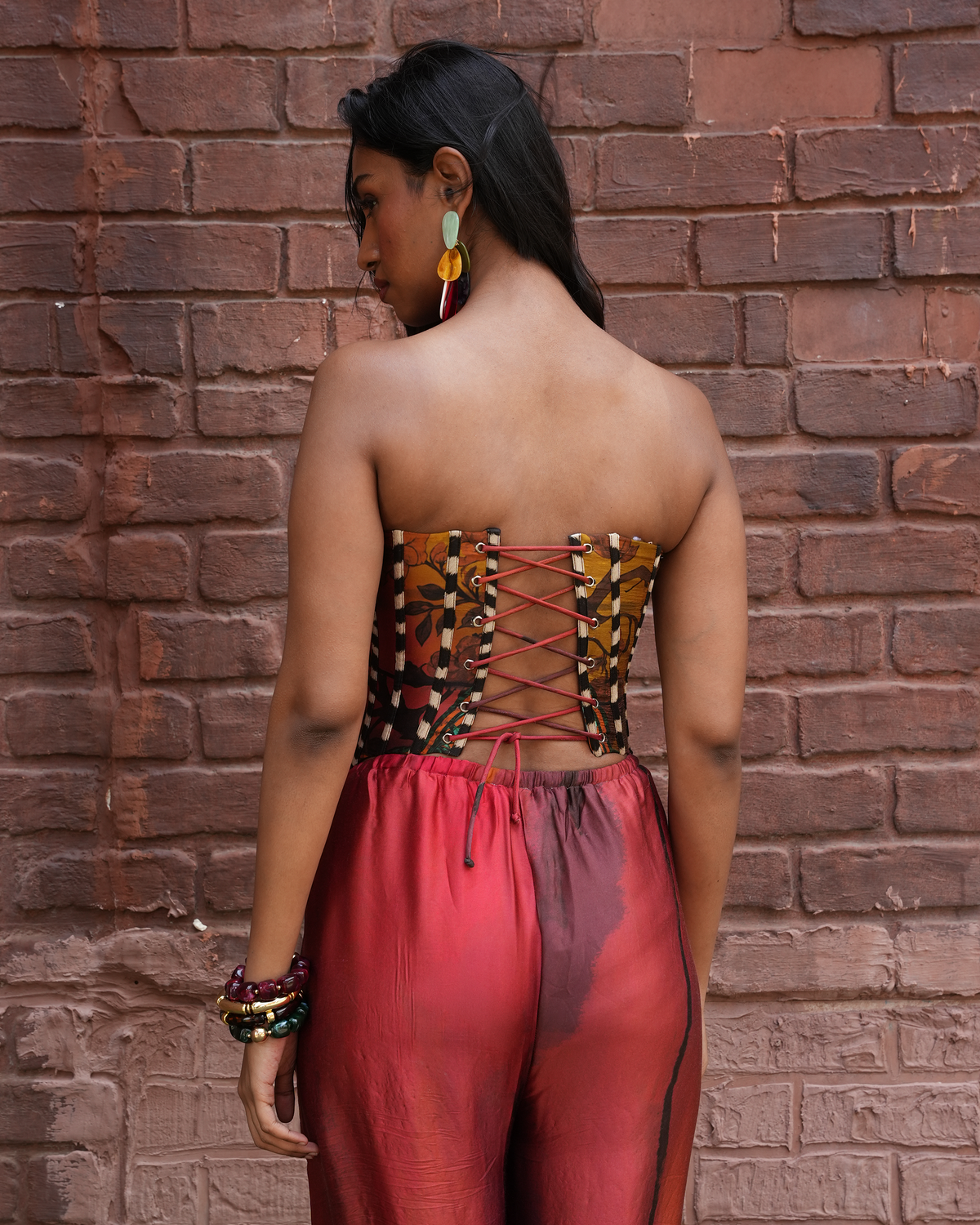 A woman stands with her back to the camera in front of a red-brown brick wall, wearing a strapless corset-style top with brown, gold, and red lace-up detailing, paired with red satin pants.