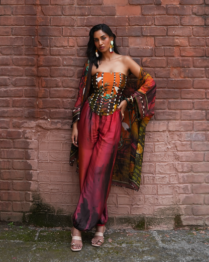 A woman models a vibrant, multicolored traditional Indian outfit consisting of a strapless top with a matching embroidered corset, wide-leg trousers in a deep red and purple hue, and a patterned dupatta, posing against a rustic red brick wall.