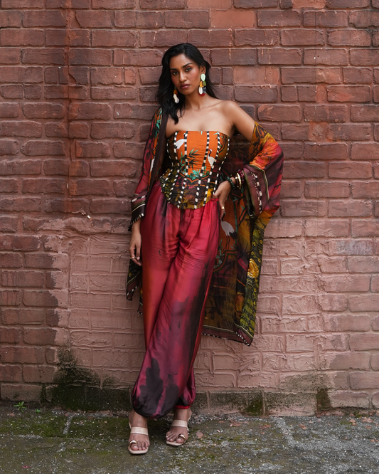 A woman models a vibrant, multicolored traditional Indian outfit consisting of a strapless top with a matching embroidered corset, wide-leg trousers in a deep red and purple hue, and a patterned dupatta, posing against a rustic red brick wall.