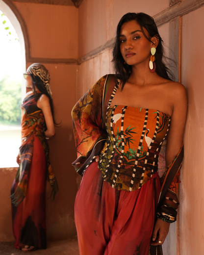 A woman models a bohemian-style printed outfit consisting of a strapless top with a bamboo motif, matching patterned sleeves, and a rust-red skirt, standing in a warm-toned corridor with her reflection visible in the background.