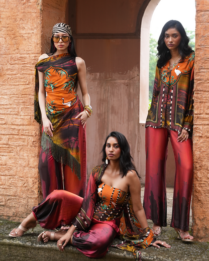 Three models showcase vibrant, earth-toned printed clothing in an outdoor stone setting with a terracotta archway. The outfits feature rich patterns of orange, red, brown, and black, with one model wearing a matching dupatta.