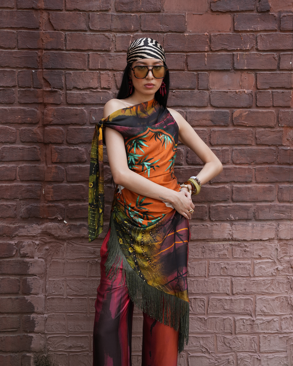 A model wearing a vibrant, multicolored tropical print scarf and matching pants stands against a rustic red-brown brick wall. The scarf features a mix of orange, green, yellow, and black patterns with a fringed hem.