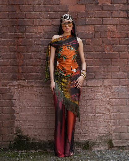 A woman stands against a red-brown brick wall wearing a one-shoulder maxi dress with a tropical print. The dress features a vibrant orange and green palm tree pattern on the upper half, a black and red gradient lower half with fringe, and a patterned headscarf and sunglasses.