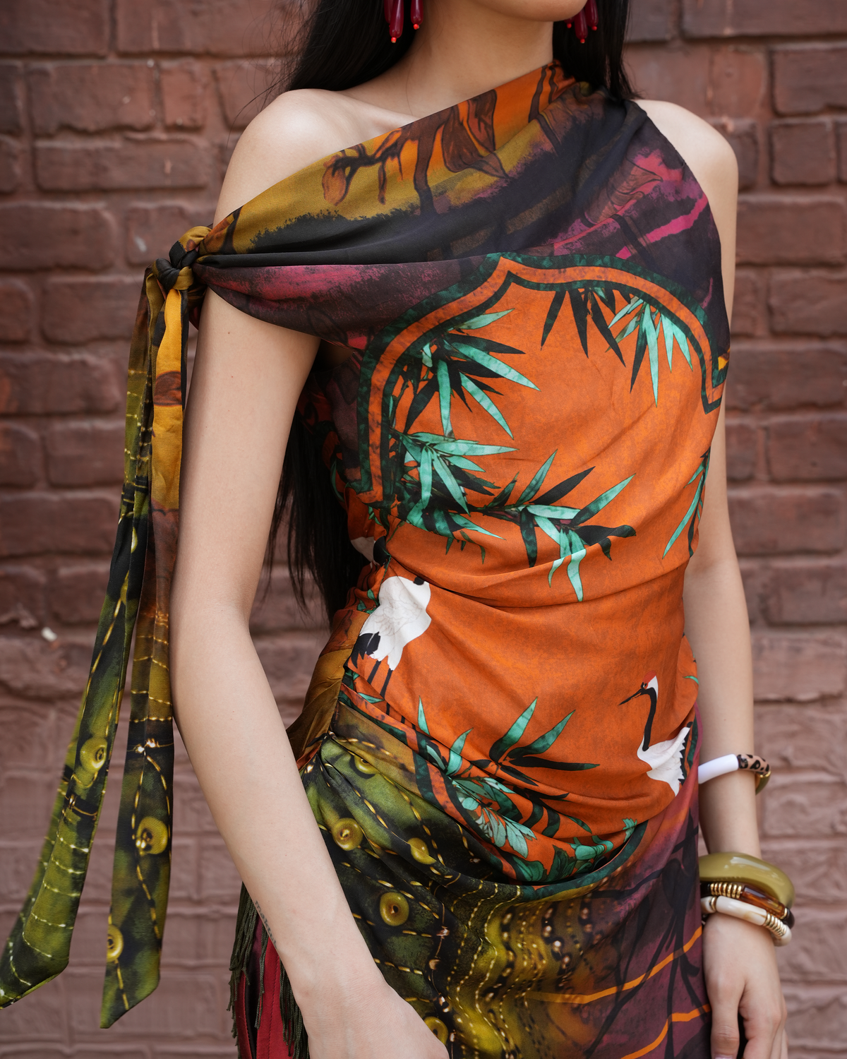 A woman wearing a one-shoulder printed scarf dress with a vibrant orange, green, black, and white pattern, tied at her shoulder, posed against a red brick wall.