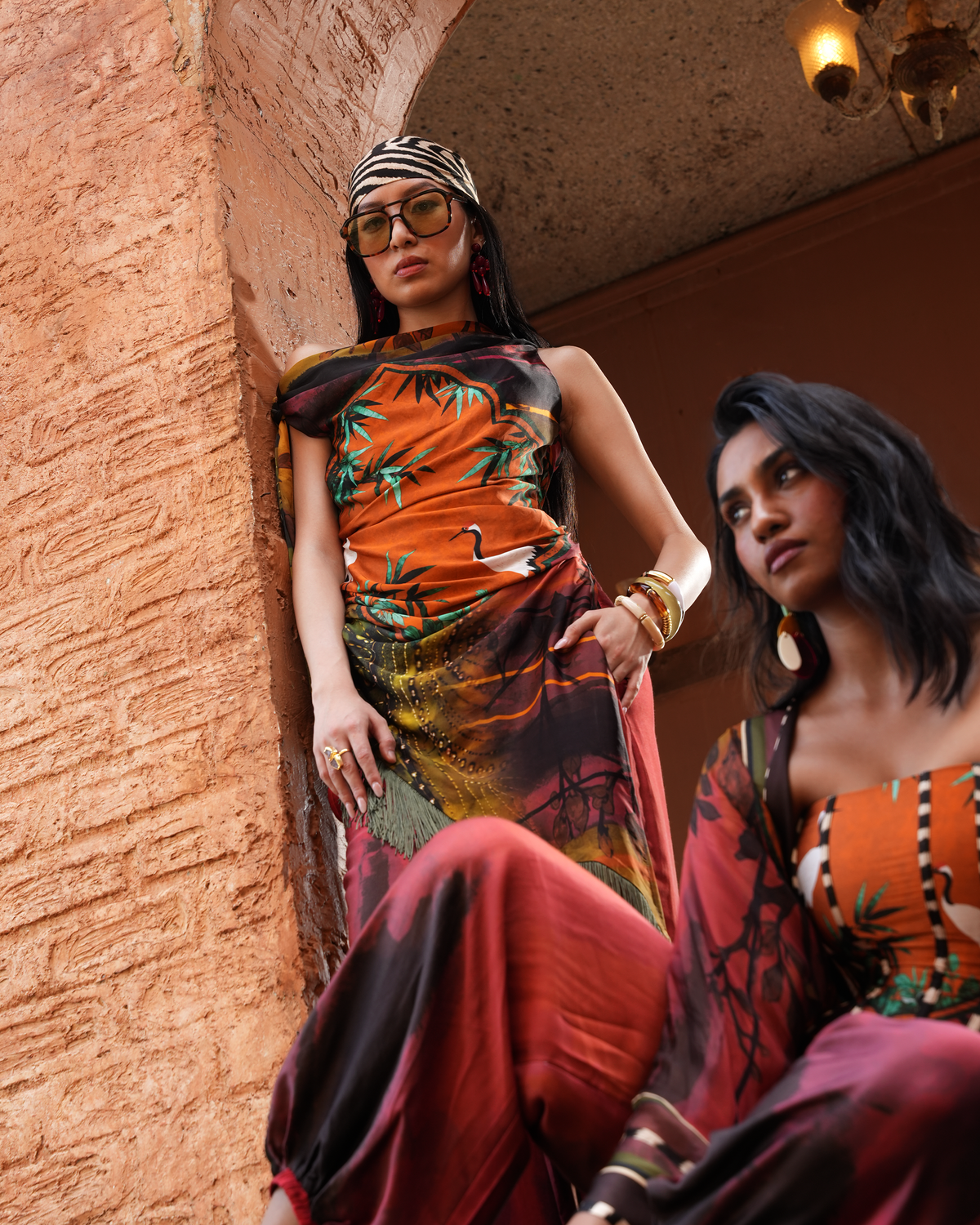 Two women modeling a vibrant orange and red tropical print outfit in a warm, rustic architectural setting with textured terracotta walls.