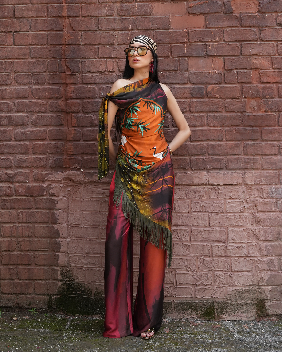 A woman wearing a colorful printed scarf and matching draped outfit stands against a red brick wall. The outfit features a vibrant orange, black, green, and red pattern with fringe details.