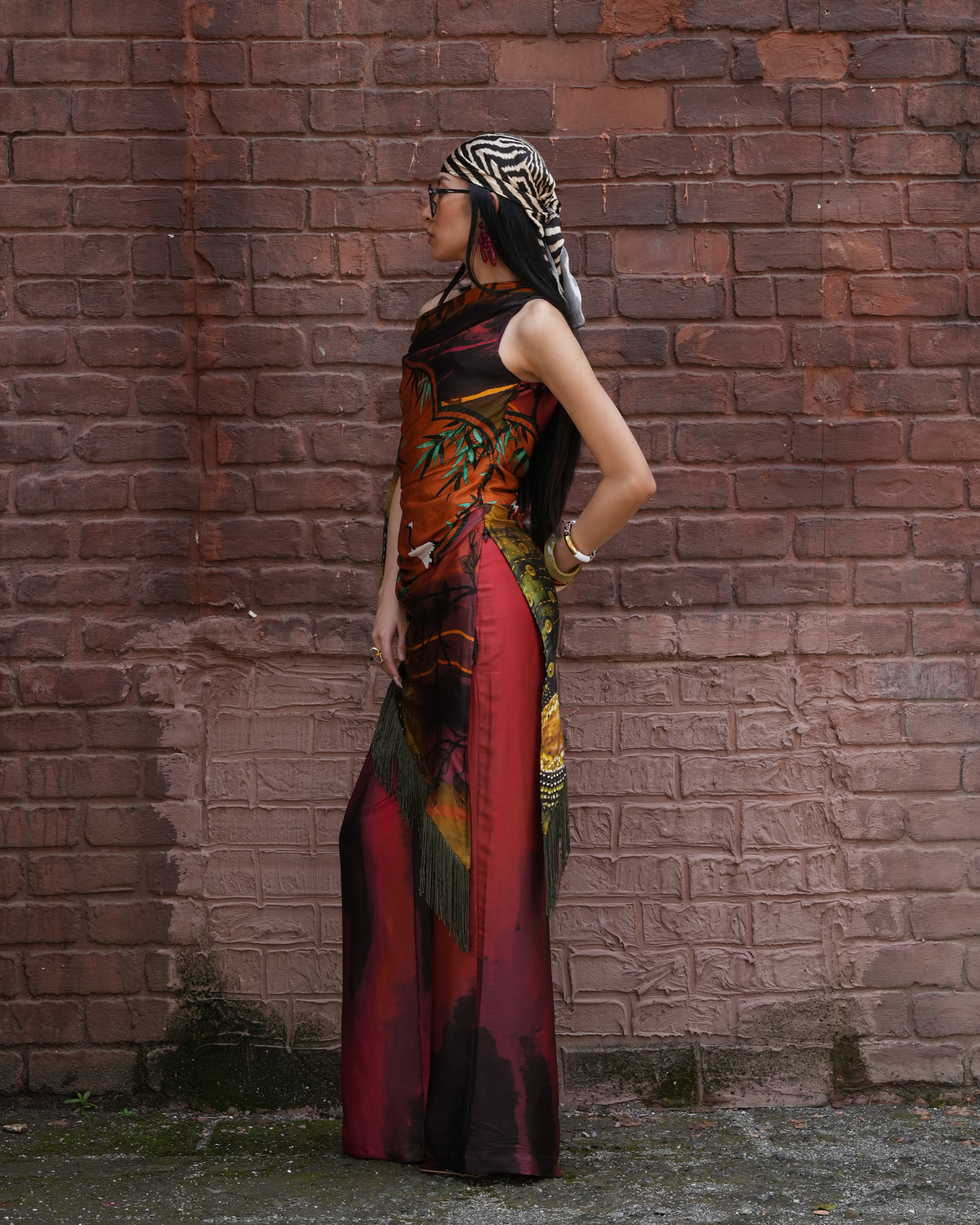 A woman stands in profile against a red-brown brick wall, wearing a sleeveless maxi dress with a bold multicolor print featuring red, orange, black, green, and yellow tones, along with a black-and-white patterned headscarf.