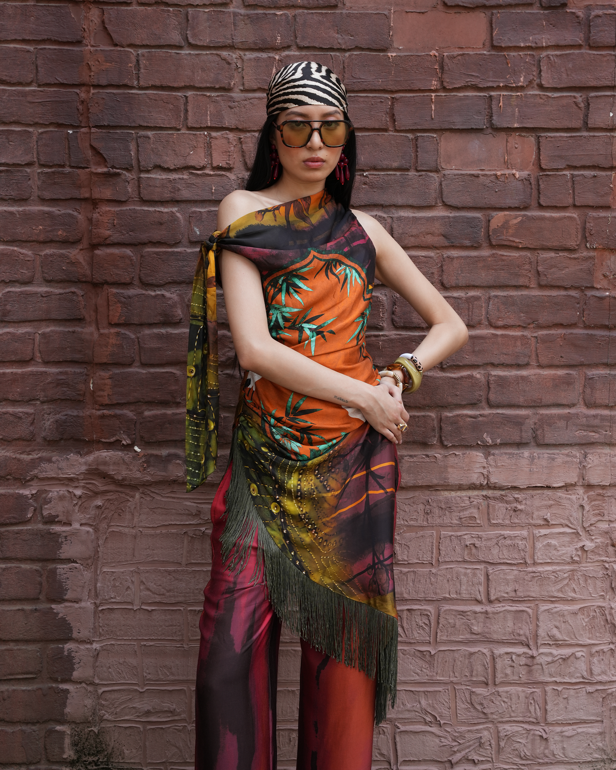 A woman stands against a red-brown brick wall wearing a colorful printed scarf draped over her shoulders and torso, paired with a black-and-white zebra-print headwrap, gold-framed sunglasses, and red pants.