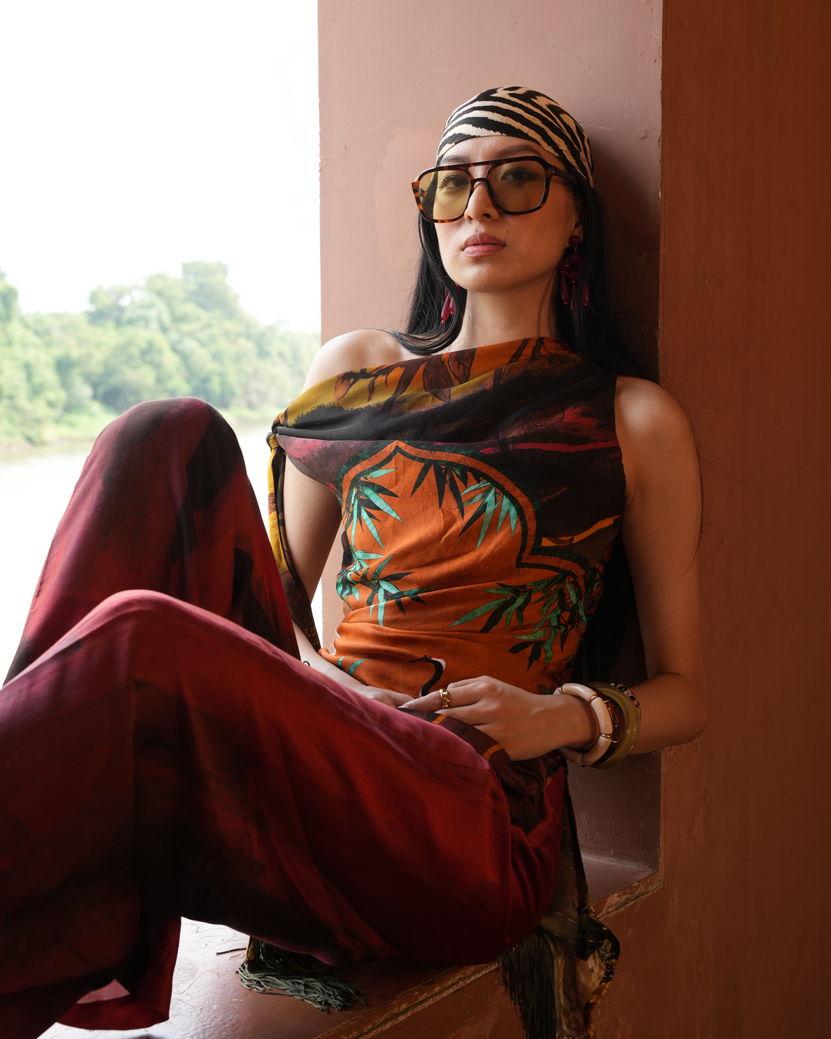 A woman wearing a stylish orange and black tropical print top, matching red pants, a tiger-print headband, and oversized sunglasses, sitting in a warm-toned architectural niche.