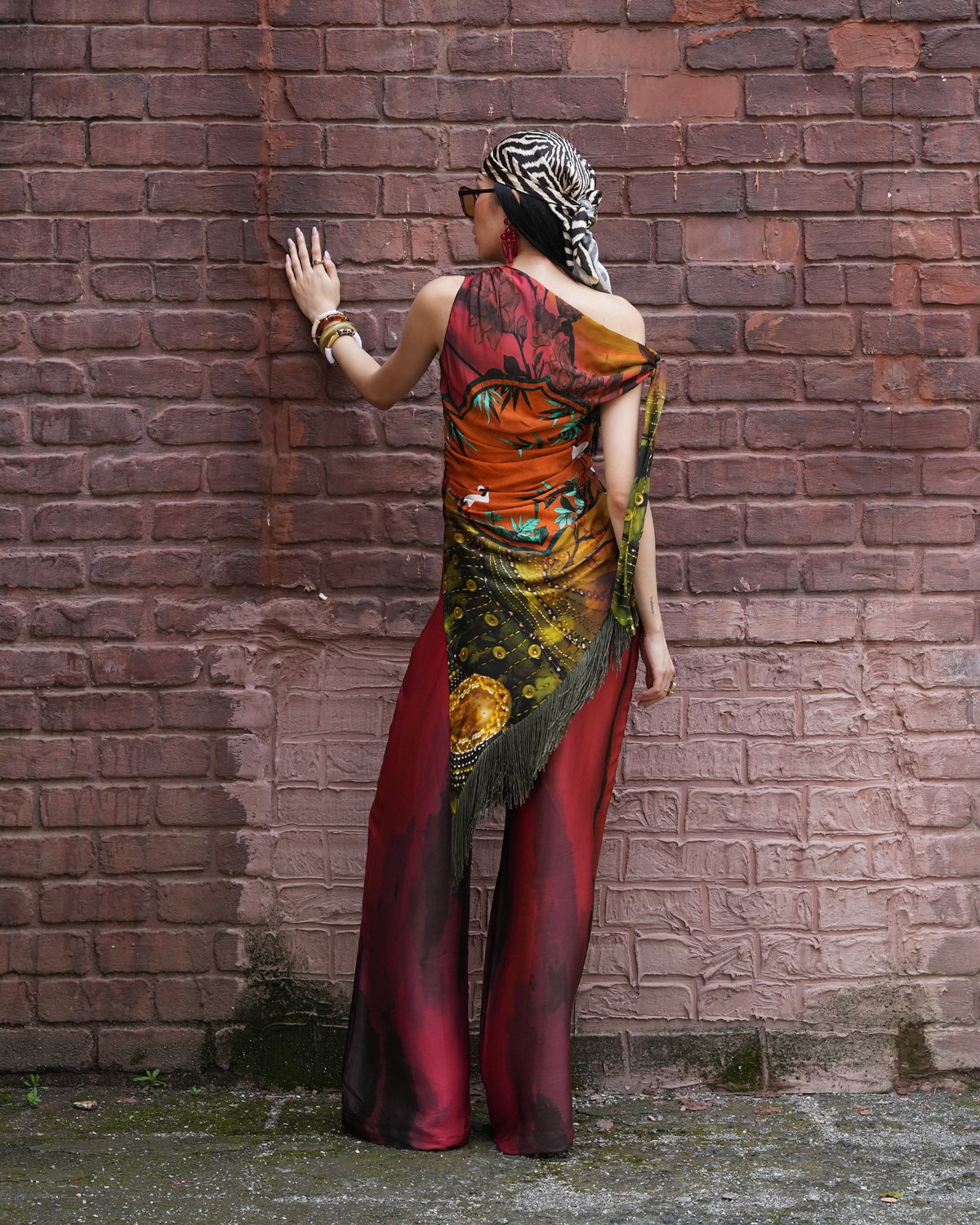 A woman stands with her back to the camera against a weathered red-brown brick wall, wearing a dramatic off-the-shoulder maxi dress with a multicolor celestial print in orange, red, black, and gold, paired with a black and white zebra-print headscarf.
