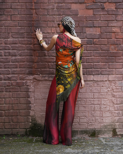 A woman stands with her back to the camera against a weathered red-brown brick wall, wearing a dramatic off-the-shoulder maxi dress with a multicolor celestial print in orange, red, black, and gold, paired with a black and white zebra-print headscarf.